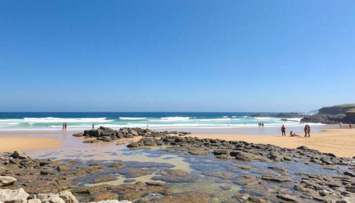 A sunny beach featuring clear waves, golden sands for sunbathing, rocky outcrops for exploration, accessible ramps, and diverse people enjoying their day under a blue sky.